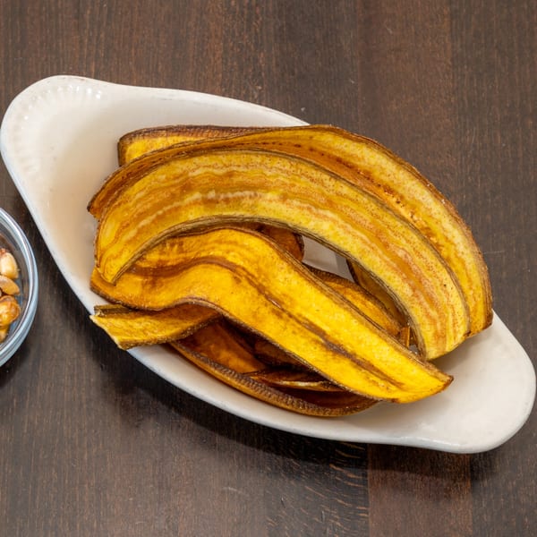 A white dish filled with golden-brown, crispy plantain chips, with a small bowl of corn in the background.