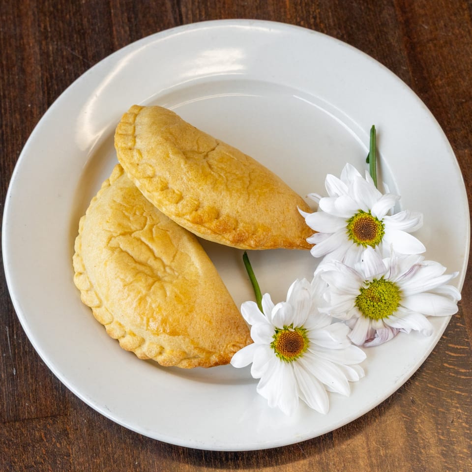 Two golden-brown Peruvian empanadas with crimped edges, garnished with white daisies on a white plate.