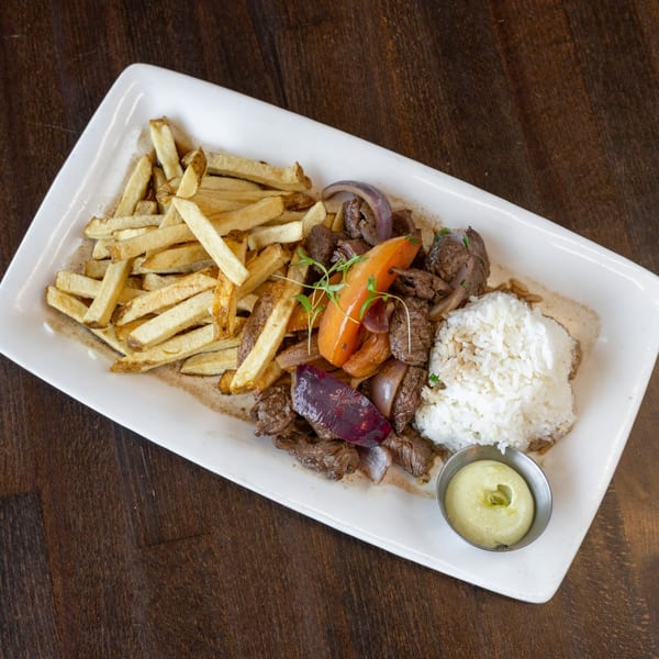 Peruvian Lomo Saltado with steak, fries, rice, onions, peppers, and green sauce on a white plate.